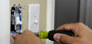 A person uses a screwdriver to install or repair a light switch on a wall next to an already installed switch.