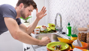 %Title% | Any Hour Services A man stands frustrated at a kitchen sink filled with dirty dishes and food scraps, with dish soap and jars on the counter.