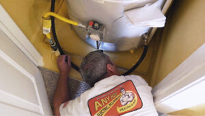 %Title% | Any Hour Services A technician in a branded shirt inspects or repairs a water heater in a small, enclosed space.