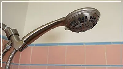 A close-up view of a metal handheld shower head mounted above pink and beige tiled walls in a bathroom.