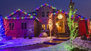 %Title% | Any Hour Services A house decorated with multicolored holiday lights, snow on the lawn, and two small trees wrapped in blue and red lights near the walkway.