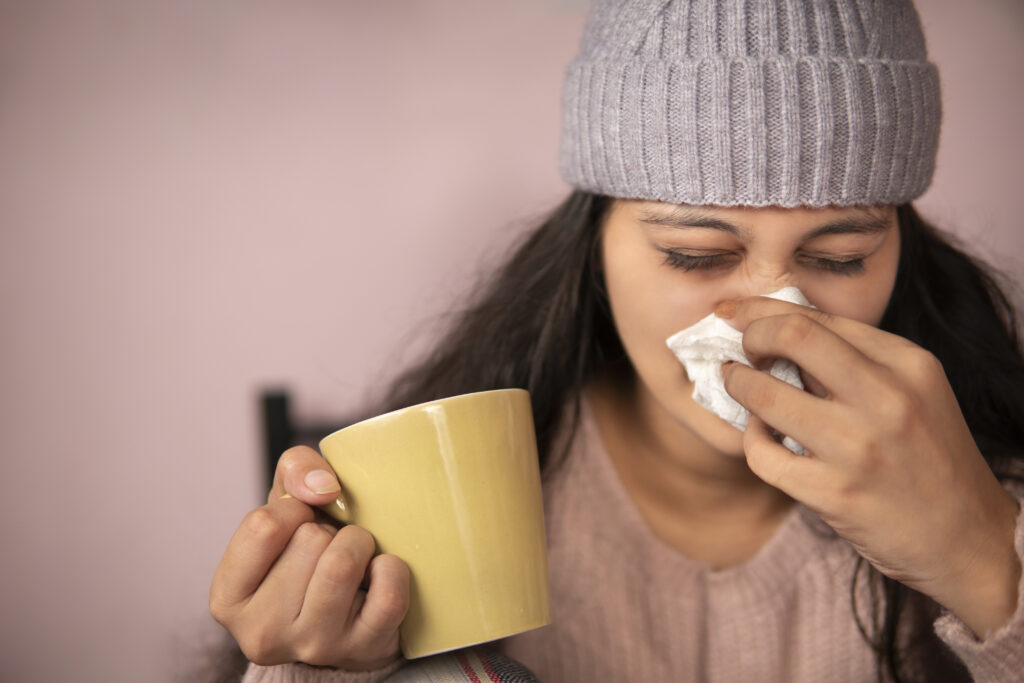 Woman wearing a knit hat holds a yellow mug in one hand and uses a tissue to blow her nose, appearing to have a cold—highlighting the need for effective indoor air quality solutions.