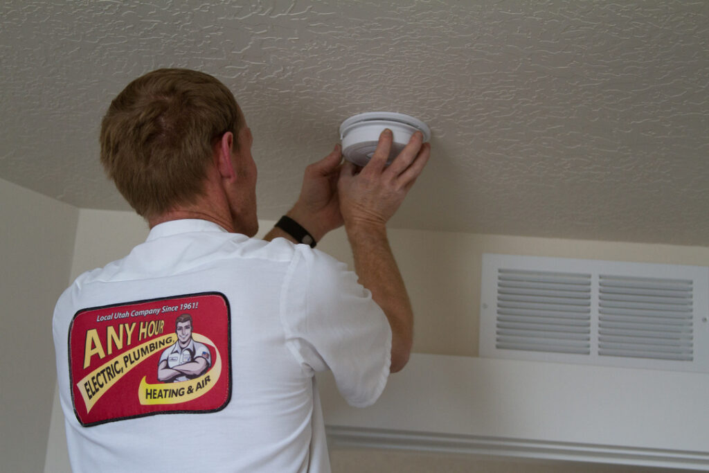 A person in a work uniform is installing or checking a smoke detector on a ceiling near an air vent.