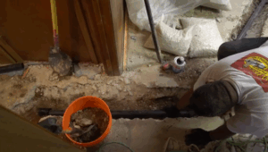 %Title% | Any Hour Services A person installs a black pipe in a trench on a concrete floor, with construction tools, a bucket, and gravel bags nearby.