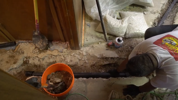 A person installs a black pipe in a trench on a concrete floor, with construction tools, a bucket, and gravel bags nearby.