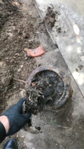 %Title% | Any Hour Services A gloved hand holds a clogged, dirty pipe covered in debris and roots, with soil and a piece of broken tile visible on the ground.
