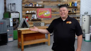 %Title% | Any Hour Services A man in a black polo shirt stands in a workshop, gesturing toward a wooden table and a triangular metal object, with tools and equipment on shelves behind him.