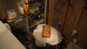 A technician in a uniform works on the lower section of an open HVAC unit in a utility room with exposed wiring and tools nearby.