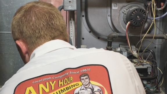 A technician in an "Any Hour Plumbing" shirt works on a furnace or HVAC system, surrounded by wires and mechanical components.