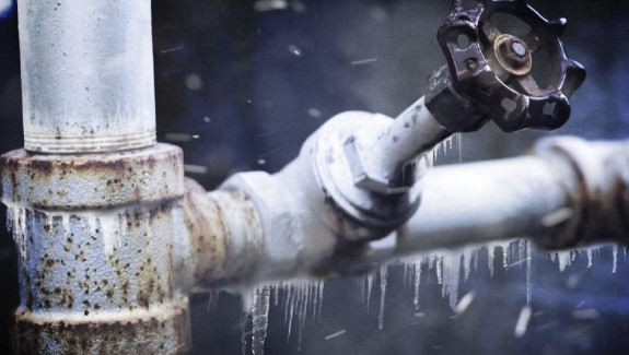 Close-up of an icy pipe with a rusted valve, surrounded by frost and icicles. The scene conveys a cold and harsh winter environment.