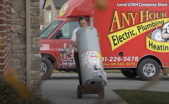 A worker walks a water heater to a suburban home from a red utility truck labeled Any Hour Electric, Plumbing, Heating & Air on a cloudy day.