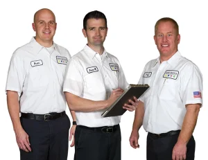 Three men in white uniforms, labeled with name tags, stand together smiling. The middle man holds a clipboard, suggesting a professional setting.