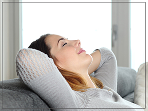 A woman in a gray sweater leans back on a sofa with her eyes closed, arms behind her head, and a peaceful expression, conveying relaxation and calm.