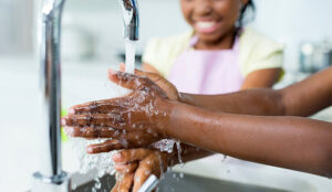 Close-up of a child washing their hands under running water in a sink, with another smiling child visible in the background.