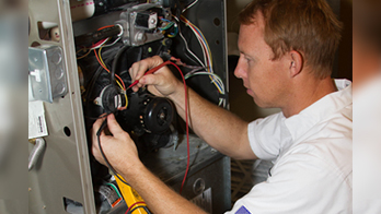 A technician in a white shirt is focused on repairing a furnace, using tools to adjust wires. The scene conveys concentration and technical skill.