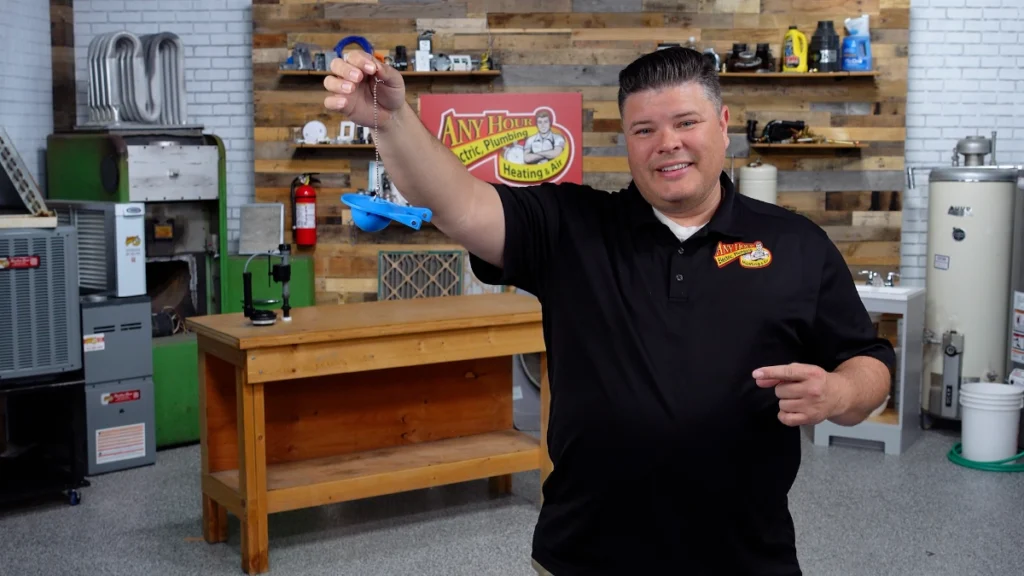 A man in a black polo shirt holds a blue plumbing tool in a workshop with shelves, tools, and equipment in the background.