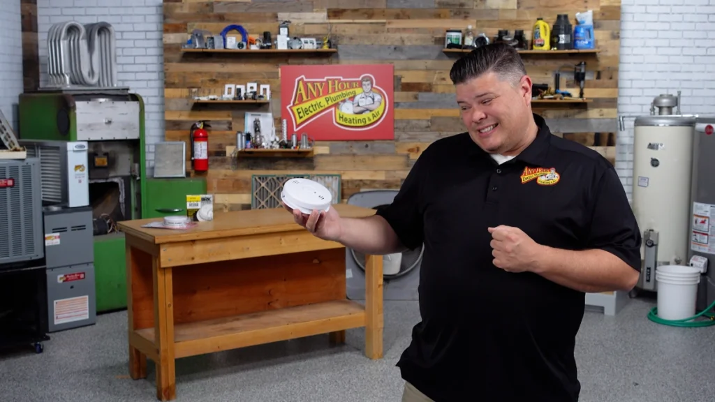 %Title% | Any Hour Services A man in a black polo shirt stands in a workshop, holding a smoke detector and smiling at the camera. A workbench and shelves with tools are visible in the background.