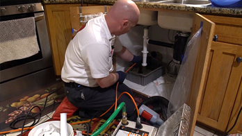 A plumber in a white shirt works under a kitchen sink, using tools and equipment. The scene conveys focus and technical expertise in plumbing repair.