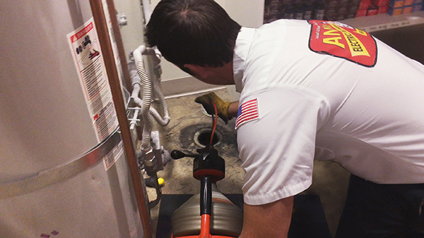 A technician in a uniform works on a plumbing issue in a utility room. He uses a tool to clear a drain near a water heater, conveying focus and expertise.