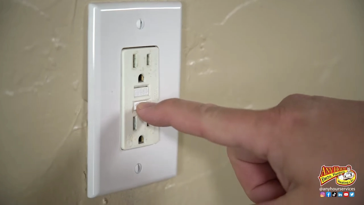 A hand presses the reset button on a white electrical outlet. The beige wall backdrop contrasts with the outlet, conveying a sense of caution.