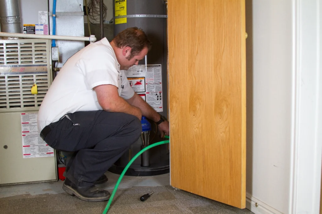 %Imagetitle% | Any Hour Services A man kneels next to a water heater, attaching a green hose to drain it, with a utility tool lying on the floor nearby.