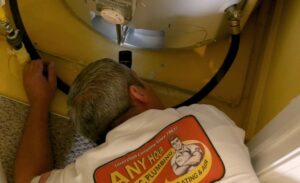 %Title% | Any Hour Services A plumber in a white "Any Hour Plumbing" shirt is inspecting pipes beneath a water heater in a small, yellow-walled space.