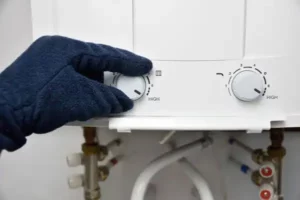 A gloved hand adjusts the temperature control dial on a white water heater, with pipes and valves visible underneath.