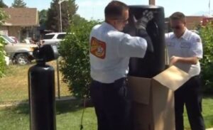 Two technicians in uniforms are unpacking a large black tank from a cardboard box outdoors, with another tank standing nearby.