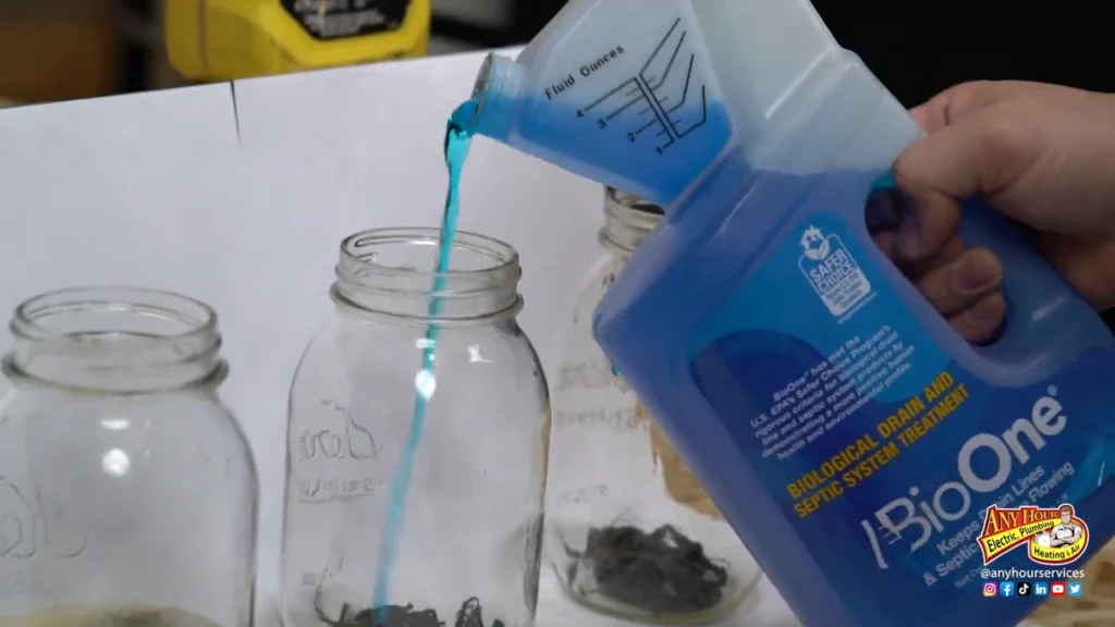 A person pours blue BioOne liquid from a measuring bottle into a clear glass jar placed among other jars on a white surface.