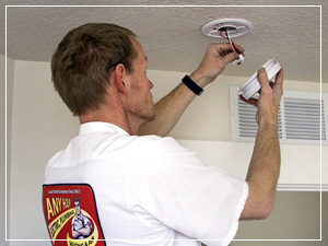 A man in a white shirt installs or repairs a smoke detector on a ceiling, holding wires and the detector base.