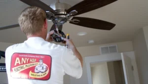 A technician in an "Any Hour Electric Plumbing Heating & Air" shirt installs or repairs a ceiling fan in a residential room.