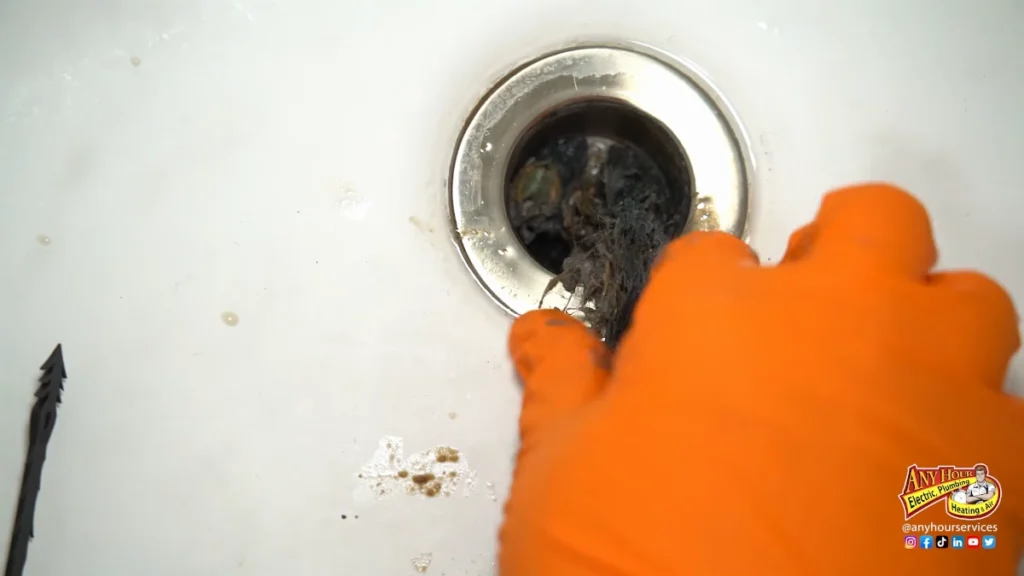 %Imagetitle% | Any Hour Services A gloved hand removes hair and debris from a sink drain, with visible grime around the drain opening.