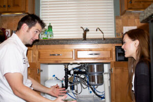 A plumber explains something to a woman while kneeling beside an open cabinet under a kitchen sink, showing exposed pipes and equipment.