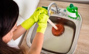 %Title% | Any Hour Services Person wearing yellow gloves uses a plunger to unclog a stainless steel kitchen sink filled with water; a bottle of dish soap is on the countertop.