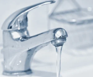 A close-up of a chrome bathroom faucet with water running from the spout. A soap dish is visible in the blurred background.