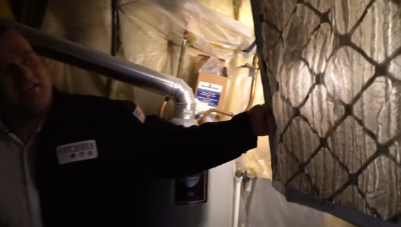 A person in a dimly lit basement holds up a used furnace filter, showing its dirtiness. The setting includes pipes and a water heater, emphasizing maintenance.