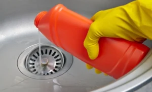 A person wearing yellow gloves pours liquid from a red bottle into a metal sink drain.