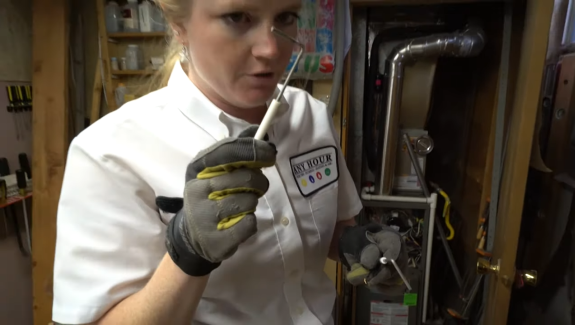 A technician in a white uniform examines a furnace part, holding a tool in a dimly lit utility room. The scene conveys focus and technical expertise.