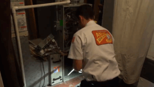 A technician in uniform is kneeling and inspecting the open panel of a furnace unit, surrounded by wires and tools in a utility room.