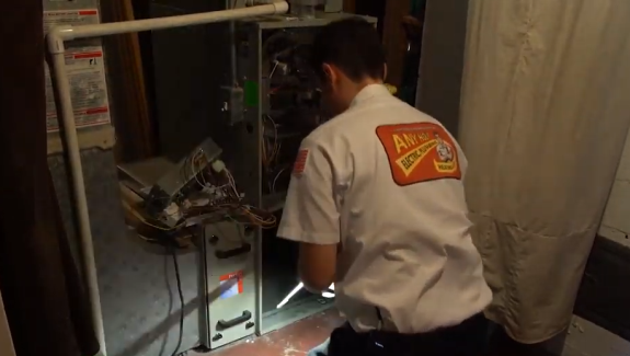 A technician in uniform is kneeling and inspecting the open panel of a furnace unit, surrounded by wires and tools in a utility room.