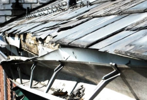 %Focuskw% | Any Hour Services A close-up view of a damaged roof with deteriorating shingles and a sagging, debris-filled rain gutter.