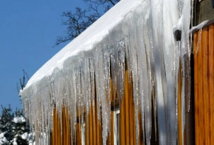 %Focuskw% | Any Hour Services Large icicles hang from the edge of a snow-covered roof on a wooden building under a clear blue sky.