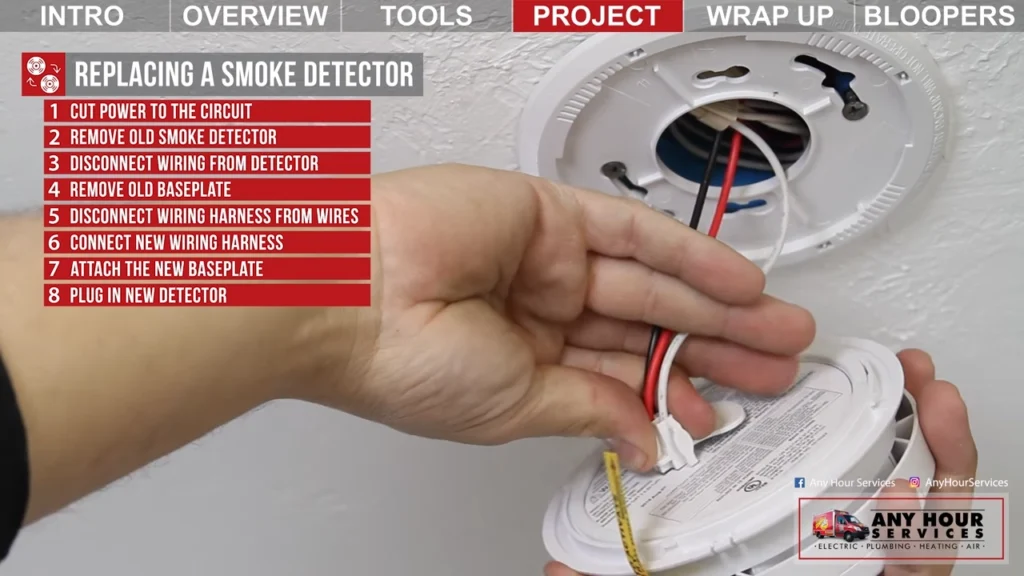 %Focuskw% | Any Hour Services A person disconnects wiring from a smoke detector mounted on a ceiling, with step-by-step replacement instructions listed on the left side of the image.