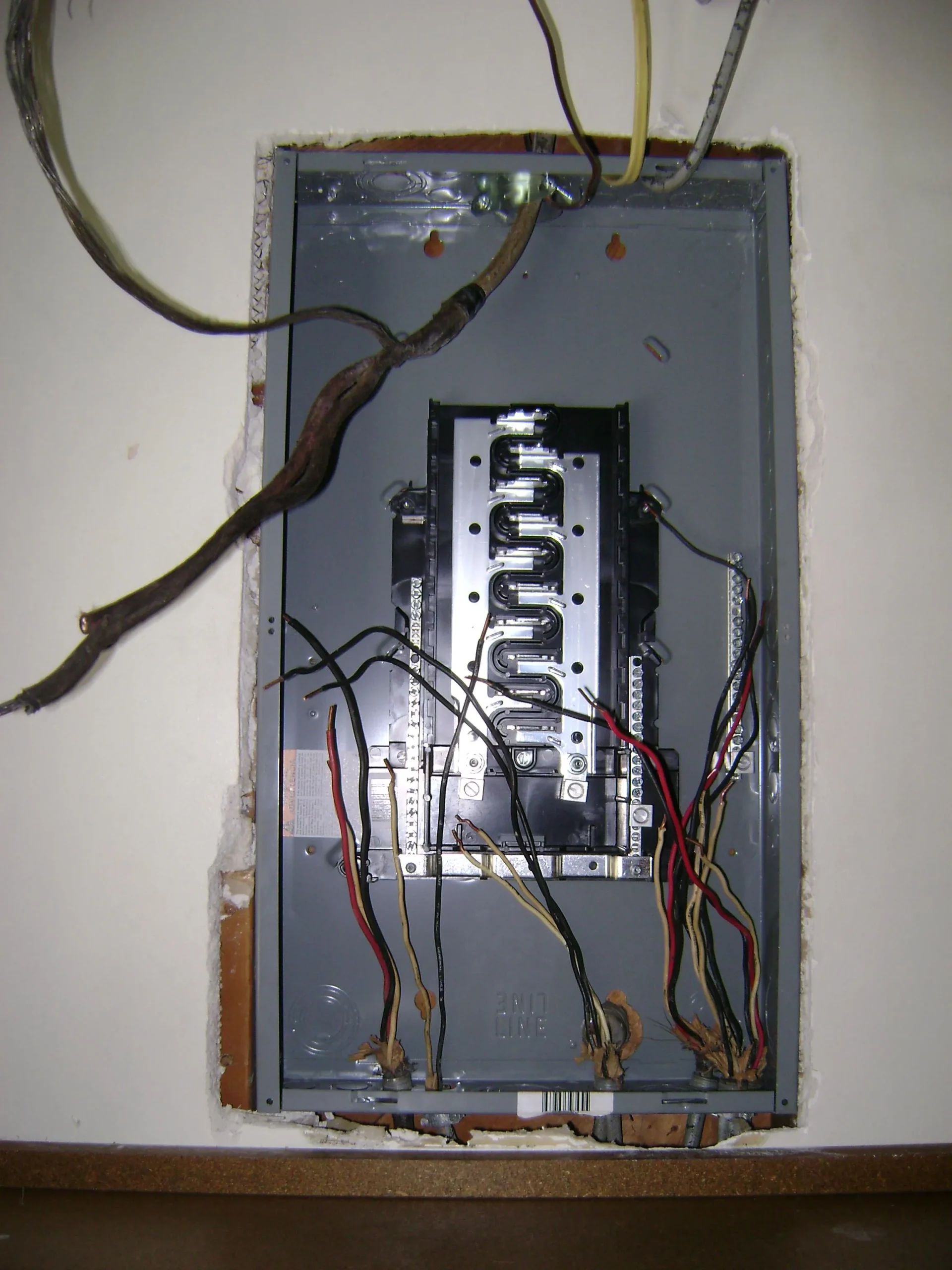 An open electrical panel installed in a wall with exposed wires and circuits connected, some insulation missing around the edges.