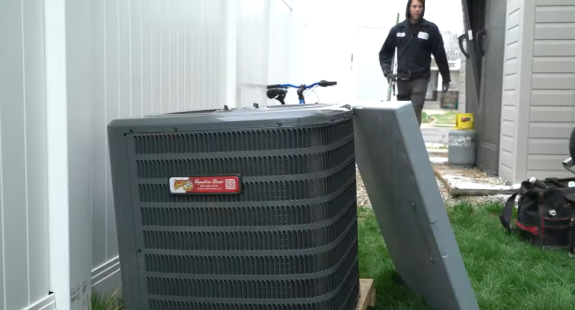 A technician approaches an outdoor air conditioning unit with its panel removed, near a white fence. Tools are visible, suggesting a repair task.