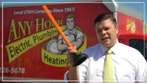 %Title% | Any Hour Services A man in a dress shirt and tie holds a plunger in front of a red "Any Hour Electric, Plumbing, Heating" service truck.