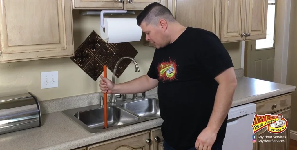 %Title% | Any Hour Services A man uses a plunger to unclog a kitchen sink while standing in front of a double basin stainless steel sink in a home kitchen.