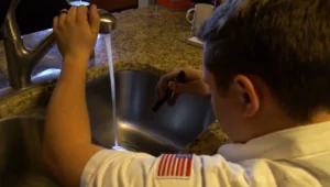 %Title% | Any Hour Services A person in a white shirt with a U.S. flag patch checks a kitchen sink under running water using a small flashlight.