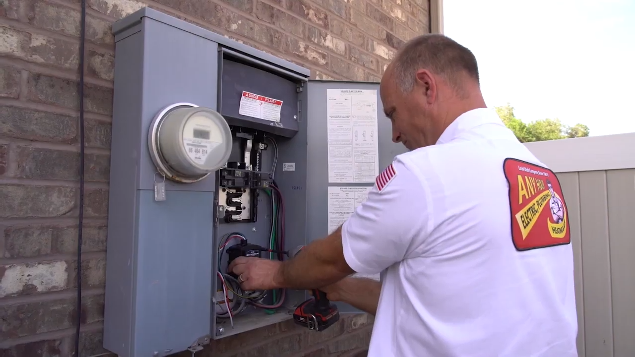 Electrician in a branded uniform works on an open electrical panel outside a brick building, using tools, focused on repairs or maintenance.
