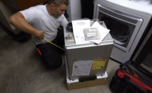 %Title% | Any Hour Services A person kneels on a concrete floor, measuring a water heater unit next to a washing machine and red tool bag, with installation manuals and parts on top of the unit.
